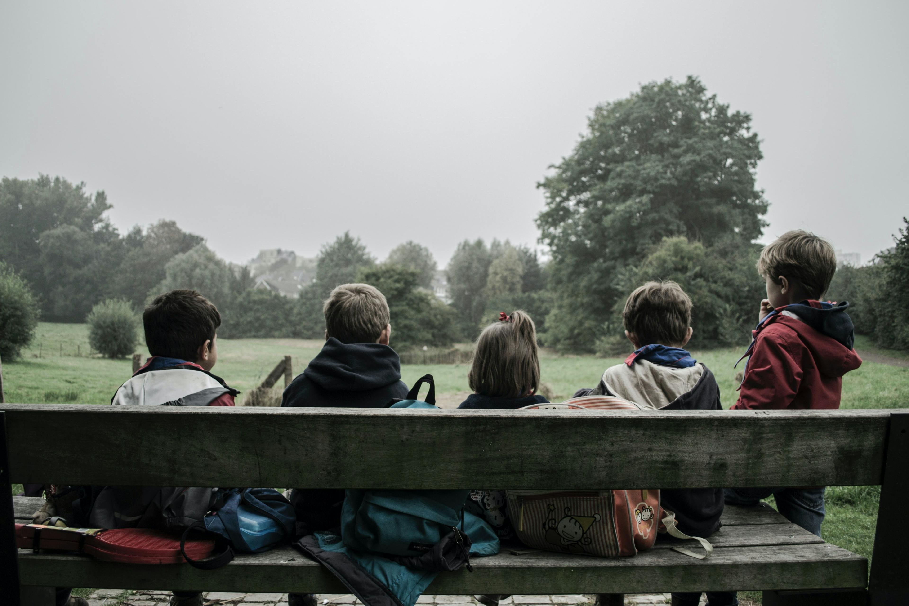 Bilde av fem barn som sitter på en benk sammen med utsikt over en stor gressplen og trær