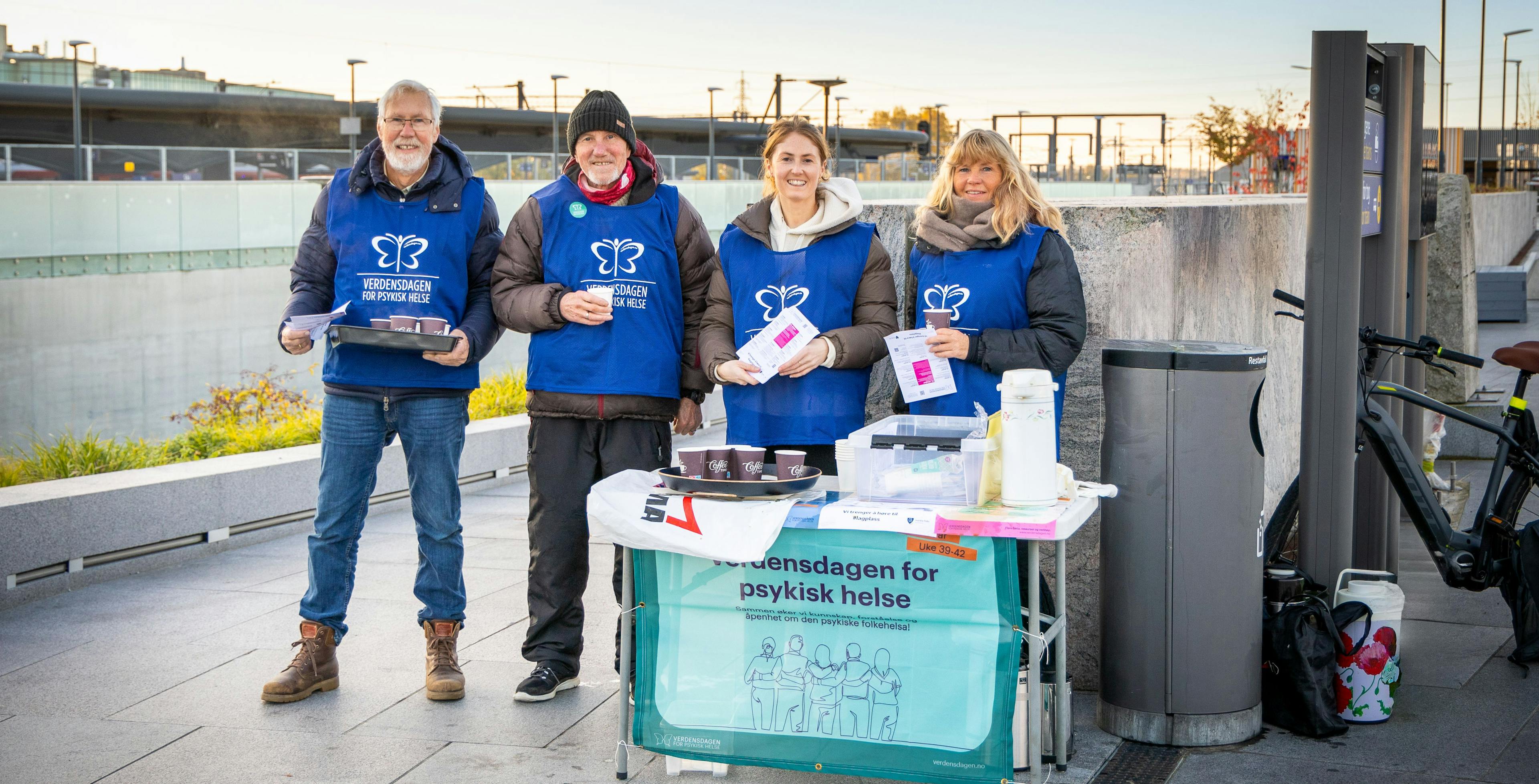 Bilde av fire smilende personer som står på en togstasjon med plakater, bannere, kaffe og vester fra Verdensdagen for psykisk helse