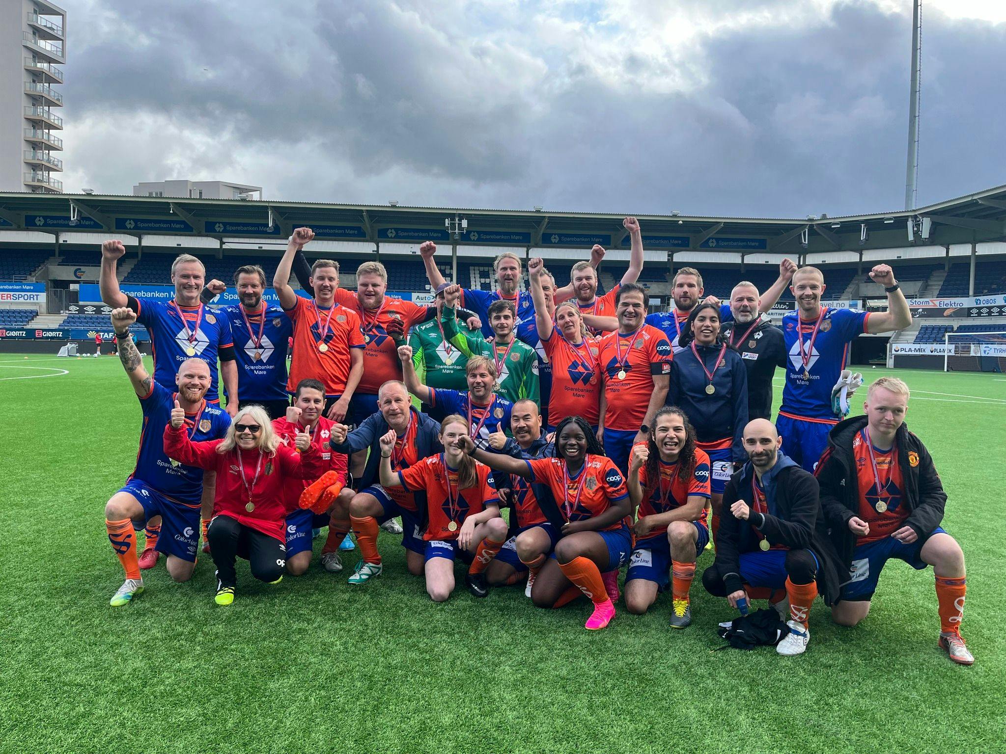 Bilde av Aalesunds fotballklubb sin markering. Bildet viser en gruppe mennesker i fotballdrakter, noen i røde, andre i blå, men alle er samlet og smiler mot kamera og løfter neven. 