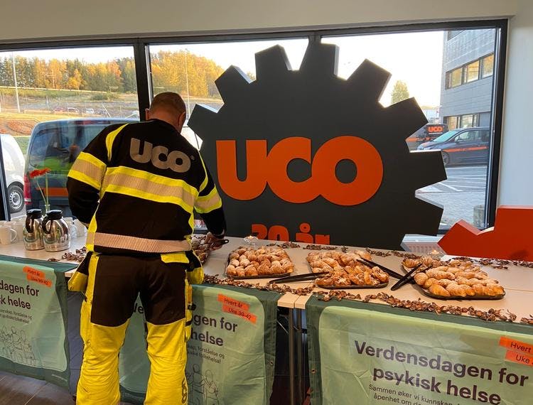 En mann i uniform med firmanavnet "UCO" printet på ryggen, står foran et bord og forsyner seg med bakst. Foran på bordene henger Verdensdagen for psykisk helse-bannere, og bak bordet står en stor plansje med nok en UCO-logo på.