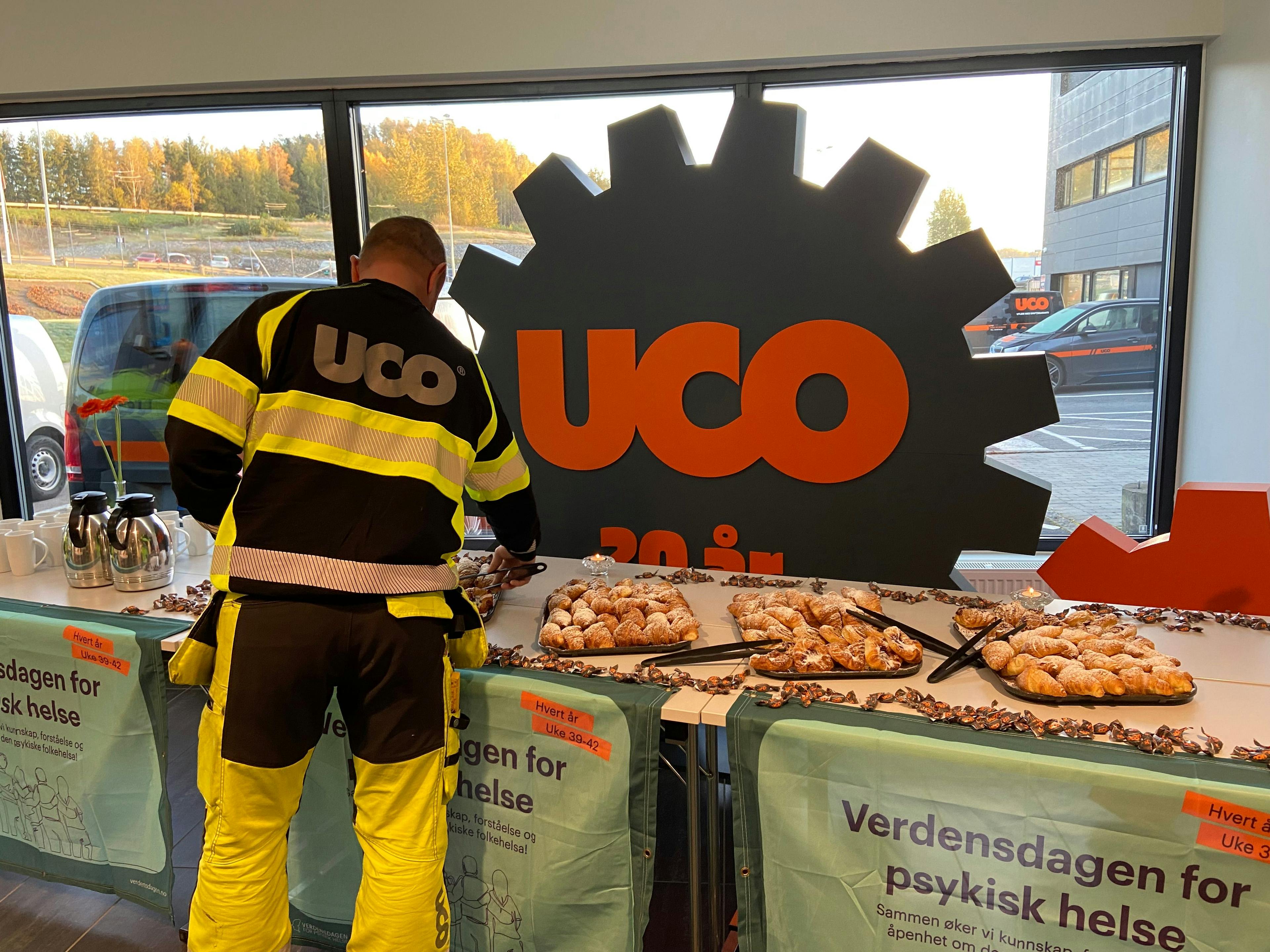 En mann i uniform med firmanavnet "UCO" printet på ryggen, står foran et bord og forsyner seg med bakst. Foran på bordene henger Verdensdagen for psykisk helse-bannere, og bak bordet står en stor plansje med nok en UCO-logo på.